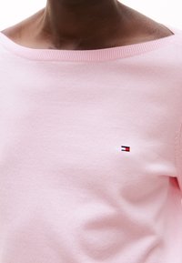 Helles rosa Sweatshirt aus weichem Stoff mit weitem Ausschnitt und geripptem Saum. Mit einem kleinen, gestickten Logo in Rot, Weiß und Blau.