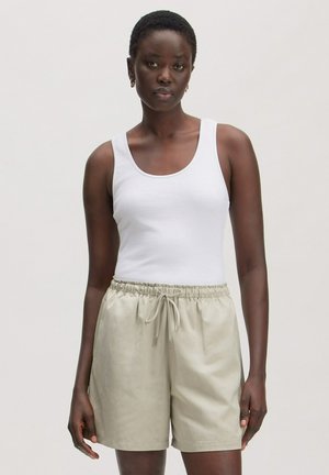 Vrouw draagt een witte mouwloze top en lichtbeige korte broek met trekkoord, staand tegen een effen lichte achtergrond.
