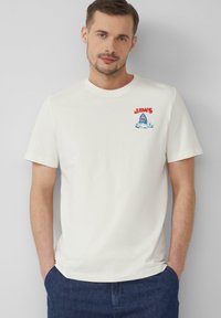T-shirt en coton blanc avec un logo rouge "JAWS" et un graphisme de requin avec des vagues bleues. Manches courtes, col rond, coupe décontractée.