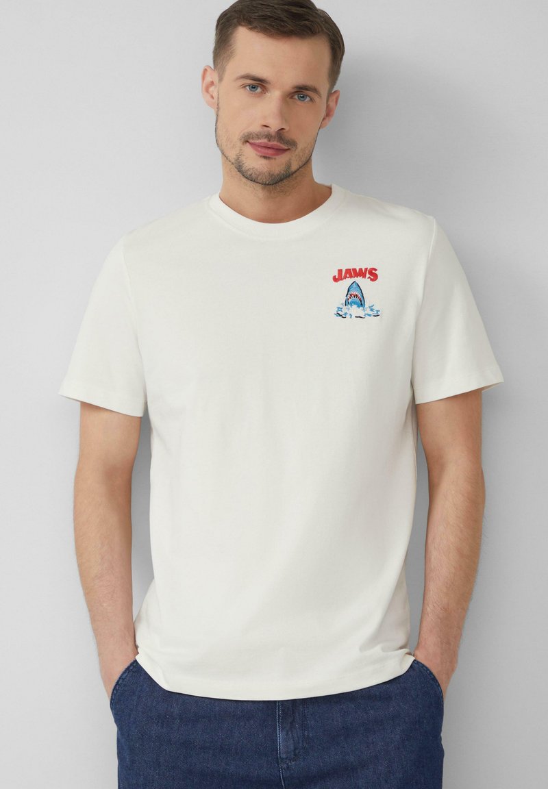 T-shirt en coton blanc avec un logo rouge "JAWS" et un graphisme de requin avec des vagues bleues. Manches courtes, col rond, coupe décontractée.