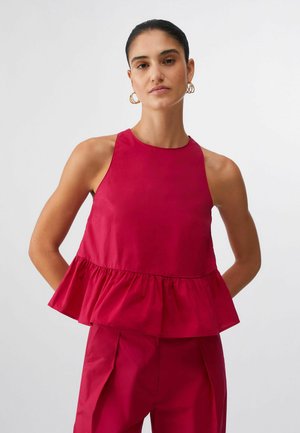 Ženska s temnimi lasmi v rdeči brezrokavnici z volančki in ujemajočih se hlačah z visokim pasom, stoji pred enostavnim svetlim ozadjem.