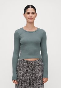 Jeune femme aux cheveux bruns portant un haut ajusté à manches longues vert et un pantalon taille haute à imprimé zèbre, se tenant devant un fond clair et uni.
