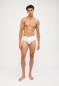 Slip da uomo bianco in cotone con una vestibilità aderente, caratterizzato da un morbido elastico in vita con dettagli del logo. Il modello è in piedi a piedi nudi, mostrando la forma e il taglio del capo.