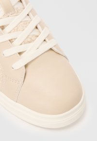 Baskets beiges avec une tige en cuir lisse, des accents texturés et des lacets blancs. Dispose d'un bout rond et d'une semelle extérieure épaisse et texturée.