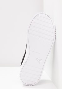 Semelle d'une basket blanche Puma avec des rainures peu profondes et le logo Puma visible au centre, appuyée contre un mur blanc.
