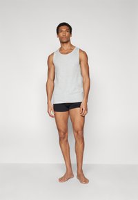 Graues Baumwoll-Top mit weiten Armausschnitten, kombiniert mit schwarzen, enganliegenden Shorts. Das Model steht barfuß vor einem neutralen Hintergrund.