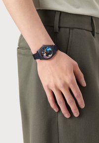 Black watch with a textured silicone strap, featuring a colorful face displaying various designs, secured on a wrist beside olive green fabric.