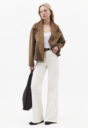 Blondynka w brązowej zamszowej kurtce, białym topie na ramiączkach, białych, szerokich spodniach z wysokim stanem, czarnym pasku, trzyma czarną torbę na ramię, patrzy na bok.
