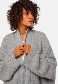 Cardigan en tricot gris avec un motif texturé, des manches larges et un col en V profond. Le vêtement est maintenu fermé à l'avant.
