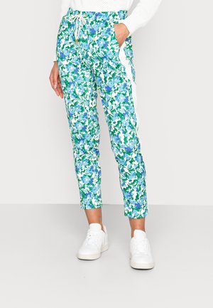 Vrouw met witte sneakers en een broek met bloemenprint in groen, blauw en wit, gecombineerd met een witte trui, staand.