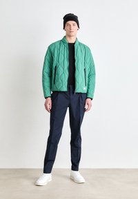 Quilted green bomber jacket with a stand collar, black zipper pockets, paired with navy pants and white sneakers. Model wears a black beanie.