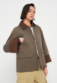 Καφέ και ελιάς πράσινο μπουφάν με γιακά από καμπάνα, φαρδιά γραμμή, δύο μπροστινές τσέπες και αντίθετες μανσέτες, φτιαγμένο από απαλό ύφασμα.