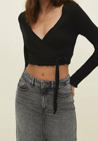 Black ribbed wrap top with long sleeves, deep V-neck, scalloped hem, and tie detail at the waist, paired with a high-waisted gray denim skirt.