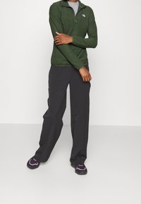 Pullover verde in fleece con colletto a zip a quarto, abbinato a pantaloni neri a gamba larga e scarpe nere da ginnastica con accenti rosa.