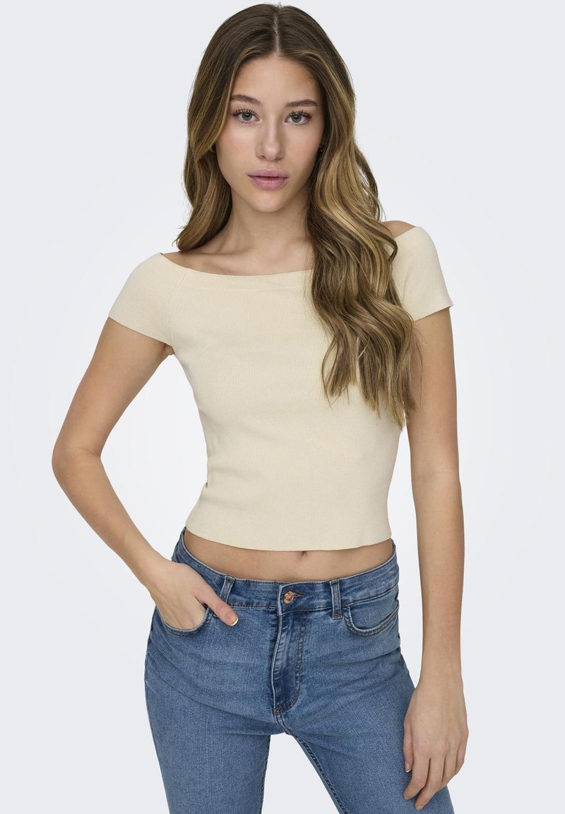 Beige ribbet crop top med off-shoulder design, korte ermer og slank passform. Kombinert med blå høyhalsede jeans med knappeåpning.