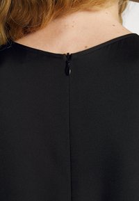 Abito nero con una texture liscia e una scollatura ampia. Presenta una zip invisibile sul retro con una piccola linguetta.