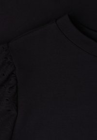 Vêtement noir avec un col rond et une manche en dentelle visible sur le côté gauche sur fond noir.