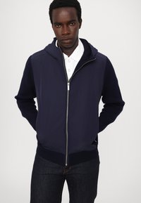 Marinblå zip-up hoodie med hög krage och ribbade ärmslut, kombinerad med en vit skjorta med krage och mörka jeans. Slät, mjuk textur.