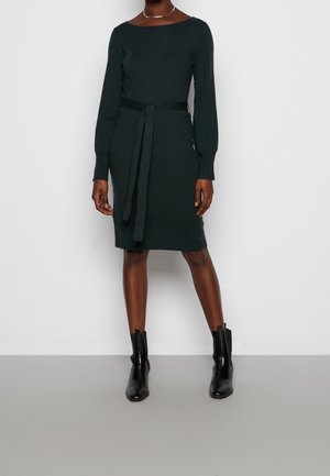 Woman standing wearing dark green long-sleeve belted knee-length dress with round neckline and black ankle boots.