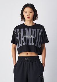 Camiseta cropped negra con gran texto gris "CHAMPION", que presenta un diseño de logotipo. Combinada con pantalones negros de cintura elástica.