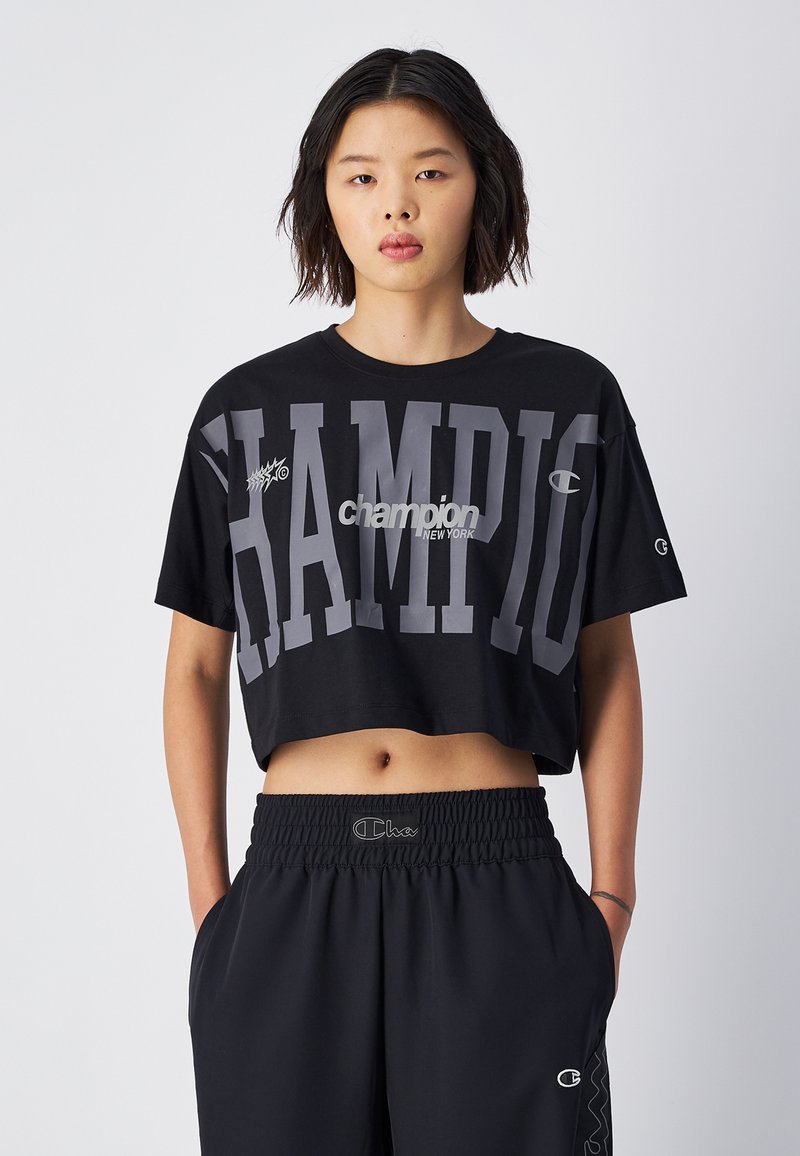 Zwarte, cropped t-shirt met grote grijze "CHAMPION" tekst, voorzien van een logo ontwerp. Gepaarde met zwarte elastische broek met tailleband.