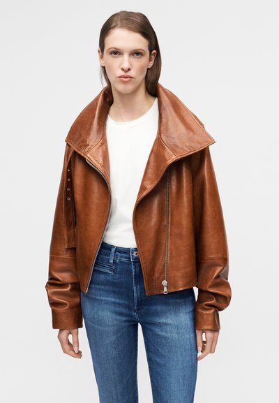 Καφέ δερμάτινο μπουφάν με ψηλό γιακά, κλείσιμο με φερμουάρ και διακριτικές ραφές. Φορεμένο πάνω από ένα λευκό πουκάμισο και μπλε τζιν.