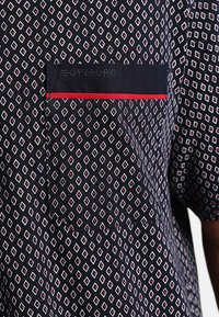 Camisa de manga corta en color navy con un patrón de diamantes en blanco y rojo. Incluye un bolsillo en el pecho con ribete rojo y un detalle de marca bordado.