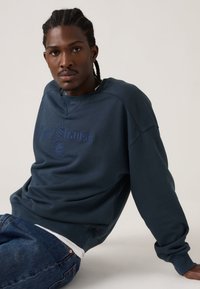 Μπλε ναυτικό φούτερ με ριμπ μανσέτες και λαιμόκοψη, κεντημένο λογότυπο σε ελαφρώς πιο σκούρα απόχρωση, σε συνδυασμό με σκούρο τζιν.