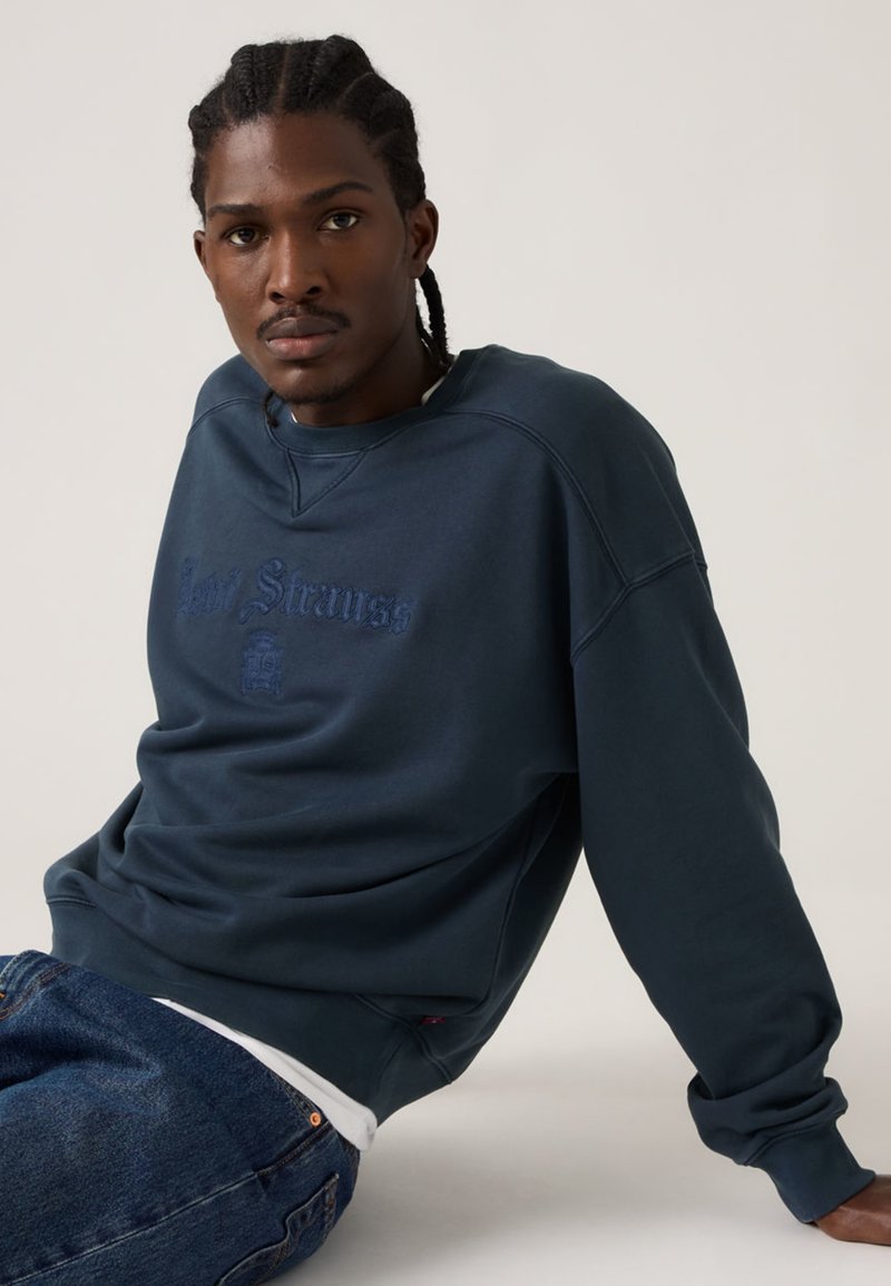 Felpa blu navy con polsini e scollo a coste, logo ricamato in una tonalità leggermente più scura, abbinata a jeans scuri in denim.