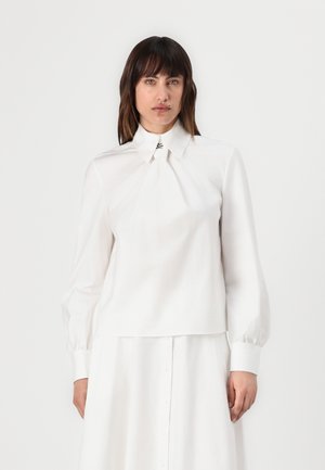 Blusa blanca de manga larga con cuello estructurado, pliegues en el frente y puños abotonados, confeccionada en un tejido suave con un acabado limpio.