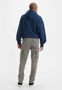 Man i marinblå hoodie och grå cargobyxor, stående med ryggen mot, framför en ljus vit bakgrund.