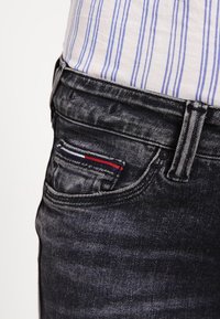 Tmavé džínsy s malou přední kapsou a červenobílými a modrými švy, doplněné světl ým pruhovaným topem pod nimi.