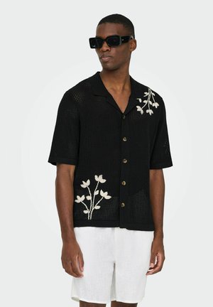 Homme portant une chemise noire à manches courtes avec des broderies florales blanches et un short blanc arrivant aux genoux, accessoirisé de grandes lunettes de soleil noires.