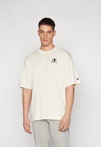 Beige Baumwoll-T-Shirt mit kurzen Ärmeln, Rundhalsausschnitt und einem kleinen schwarzen Logo auf der linken Brust. Der Stoff wirkt weich, unstrukturiert.