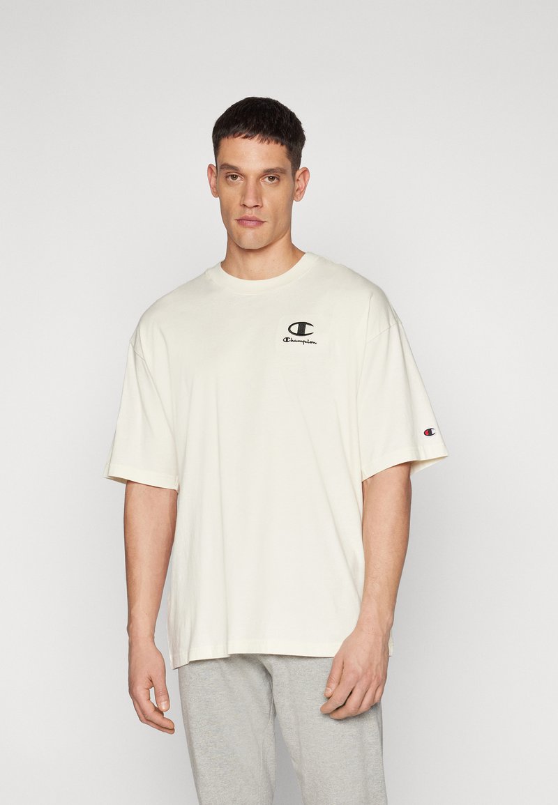 Beige Baumwoll-T-Shirt mit kurzen Ärmeln, Rundhalsausschnitt und einem kleinen schwarzen Logo auf der linken Brust. Der Stoff wirkt weich, unstrukturiert.