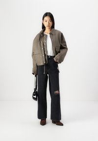 Giacca cropped verde oliva con accessori in argento, indossata sopra un maglione grigio e jeans a gamba larga neri con un ginocchio strappato. Borsa nera.