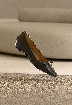 Chaussure plate en cuir noir à bout pointu avec un petit nœud décoratif et un accent doré, flottant au-dessus d'un tapis beige avec un canapé en arrière-plan.