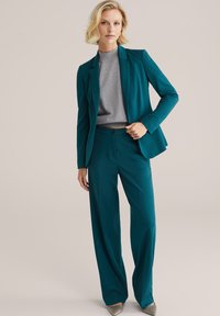 Tyrkysový blazer a odpovídající široké kalhoty vyrobené z hladké tkaniny. Pod tím je šedý přiléhavý top. Obuv je stříbrná, což dodává jemný kontrast.
