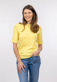 Žlutý top s krátkým rukávem a kulatým výstřihem, spárovaný se světle modrými džíny. Tkanina vypadá hladce a má volný střih.