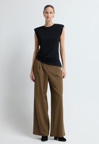 Černý top bez rukávů s drapovaným designem, kombinovaný s hnědými kalhotami s vysokým pasem a širokými nohavicemi. Oblečení má hladké textury a precizní linie.