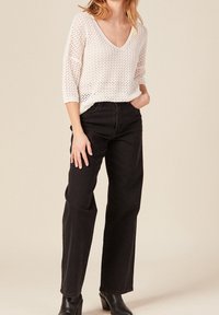 Bílý volný pletený svetr s výstřihem do V, v kombinaci s černými džíny s vysokým pasem a kotníkovými botami. Oblečení má uvolněný střih a texturované detaily.