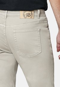 Jeans de mezclilla beige con un diseño clásico de cinco bolsillos, parche trasero con logo y detalles de costura sutiles en los bolsillos.
