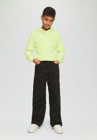 Felle gele hoodie met een kangoeroezak, gecombineerd met loszittende zwarte broek en witte sneakers. De outfit heeft een casual, moderne uitstraling.