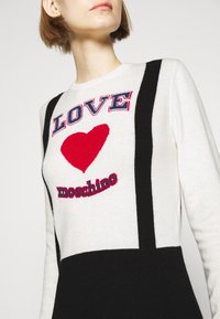 Personne portant un pull blanc et noir avec un cœur rouge et le texte "LOVE moschino" sur la poitrine.