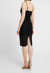 Černé krajkové šaty s přiléhavým střihem, tenkými ramínky a rozparkem až ke stehnům na zadní straně. Materiál má složité květinové vzory.