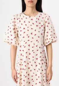 Crème jurk met bloemenpatroon met kleine rode bloemen. Volle mouwen en een ronde halslijn. Textuurstof met een subtiel plooienontwerp.