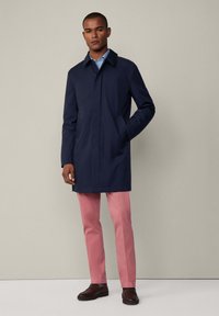 Marineblauer wasserdichter Mantel mit Umlegekragen, seitlichen Taschen, knielanger Länge, kombiniert mit rosa Hosen und braunen Slippern.