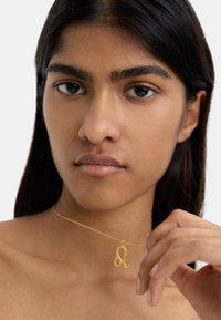 Złoty naszyjnik z stylizowanym symbolem Lwa zodiaku na cienkim łańcuszku. Zbliżenie na modelkę, która podkreśla naszyjnik na neutralnym tle.