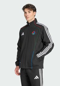 Sort atletisk jakke med lynlås foran, med hvide striber på ærmerne, farverig piping og et logo på brystet. Polyester materiale.