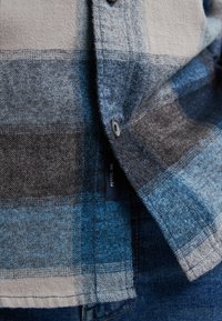 Cămașă din flanel cu carouri, în nuanțe de albastru, gri și bej; include un etichetă și detalii cu nasturi metalici. Textură moale, croială regulată.